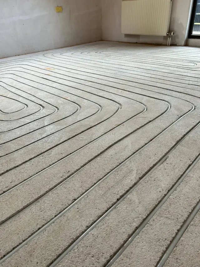 Gefräste Estrich-Nuten mit eingelegten Heizrohren einer Fußbodenheizung in einem renovierten Raum - Fenster und Heizkörper an der Wand.